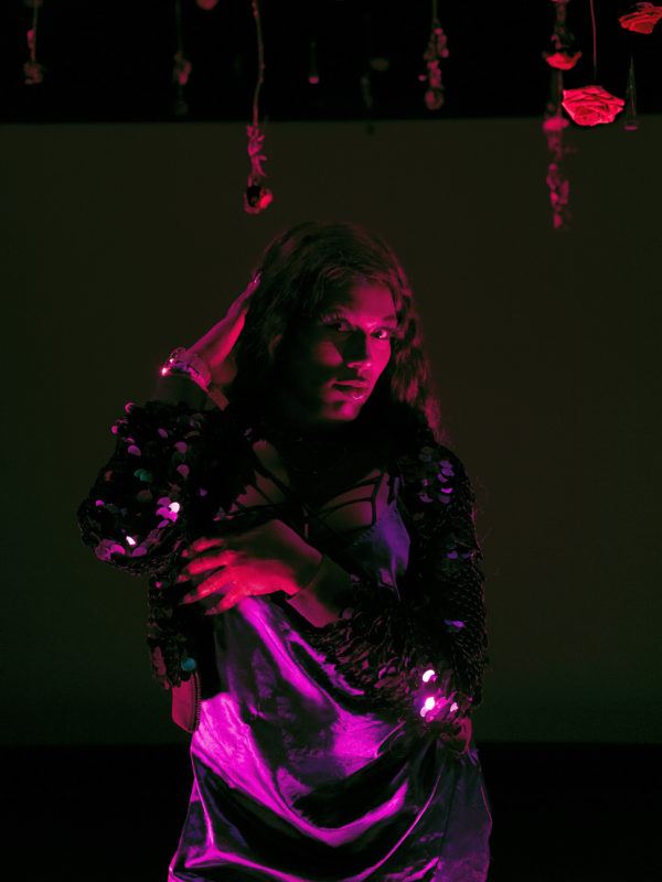 Lexii Foxx stands facing the camera in a red light, her arms posed across her chest and in her hair. She looks intently into the camera.