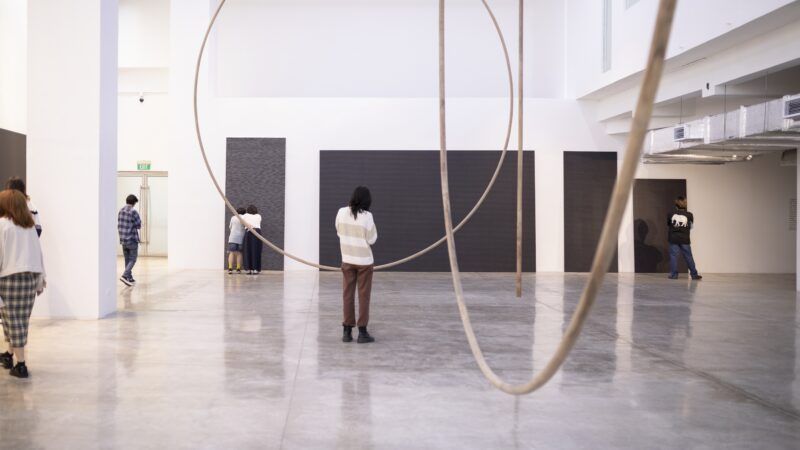 People stand and observe large black panels on the walls of a spacious, white-walled gallery with circular hanging sculptures.