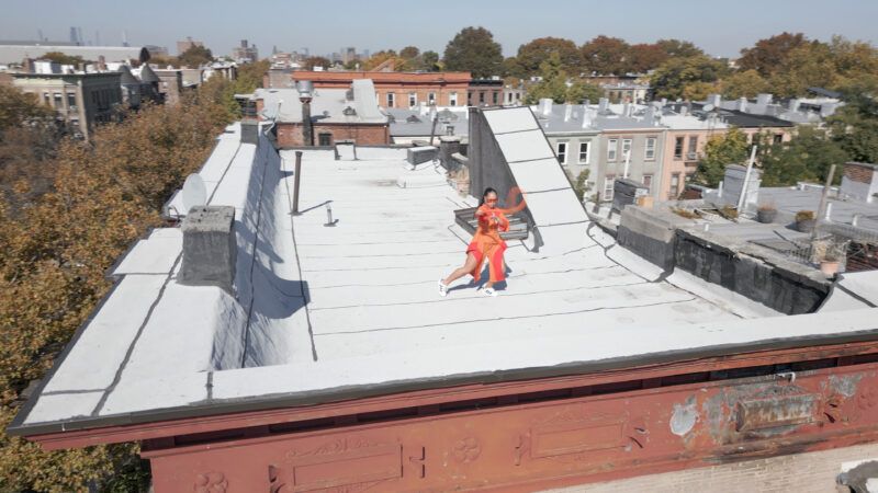 Caroline Garcia in an orange outfit stands on a flat rooftop in a residential neighborhood, surrounded by trees and buildings under a clear sky.