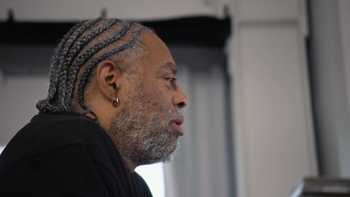 Arthur Jafa with gray braided hair, a beard, and earrings is shown in profile, looking forward in an indoor setting.