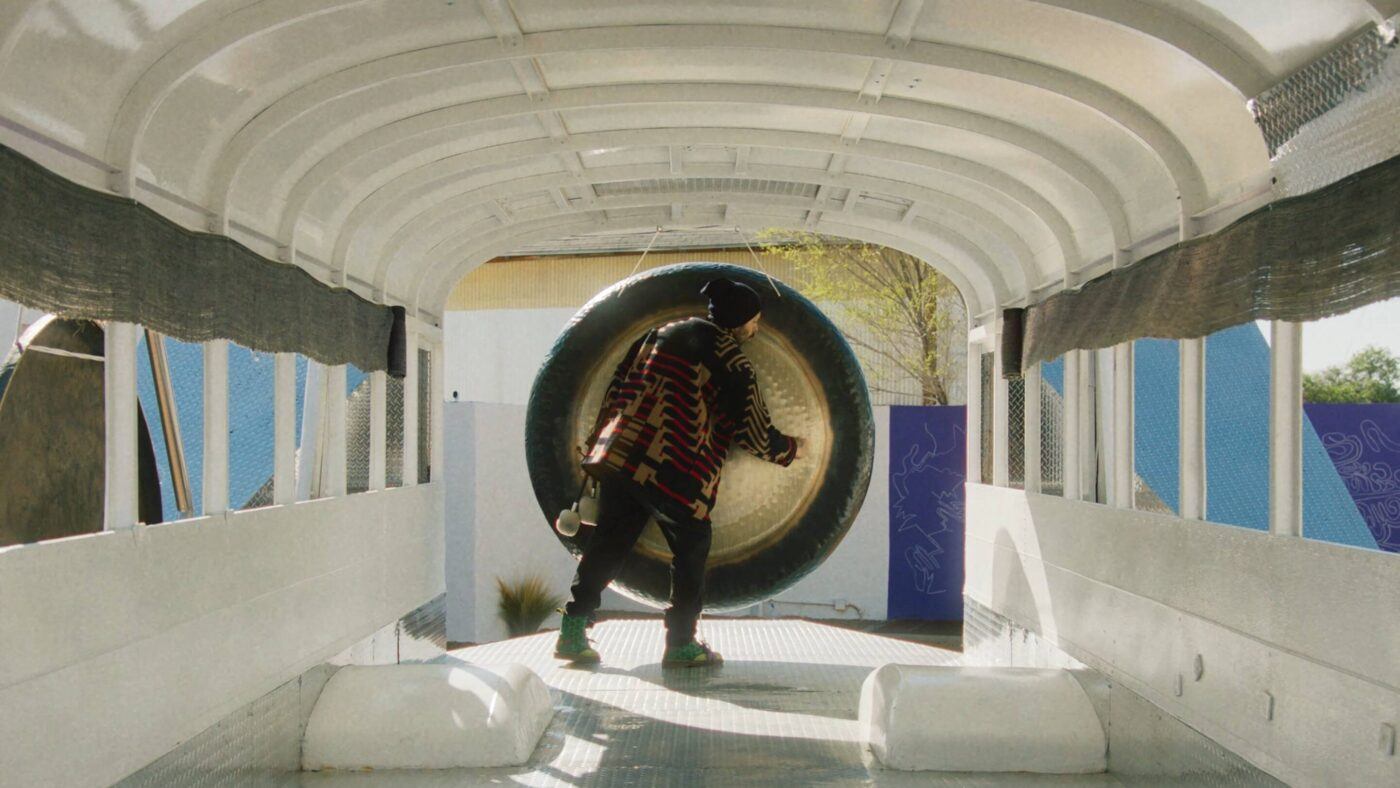 Guadalupe Maravilla with a patterned jacket and hat stands inside a white empty bus, reaching towards a large metallic gong.