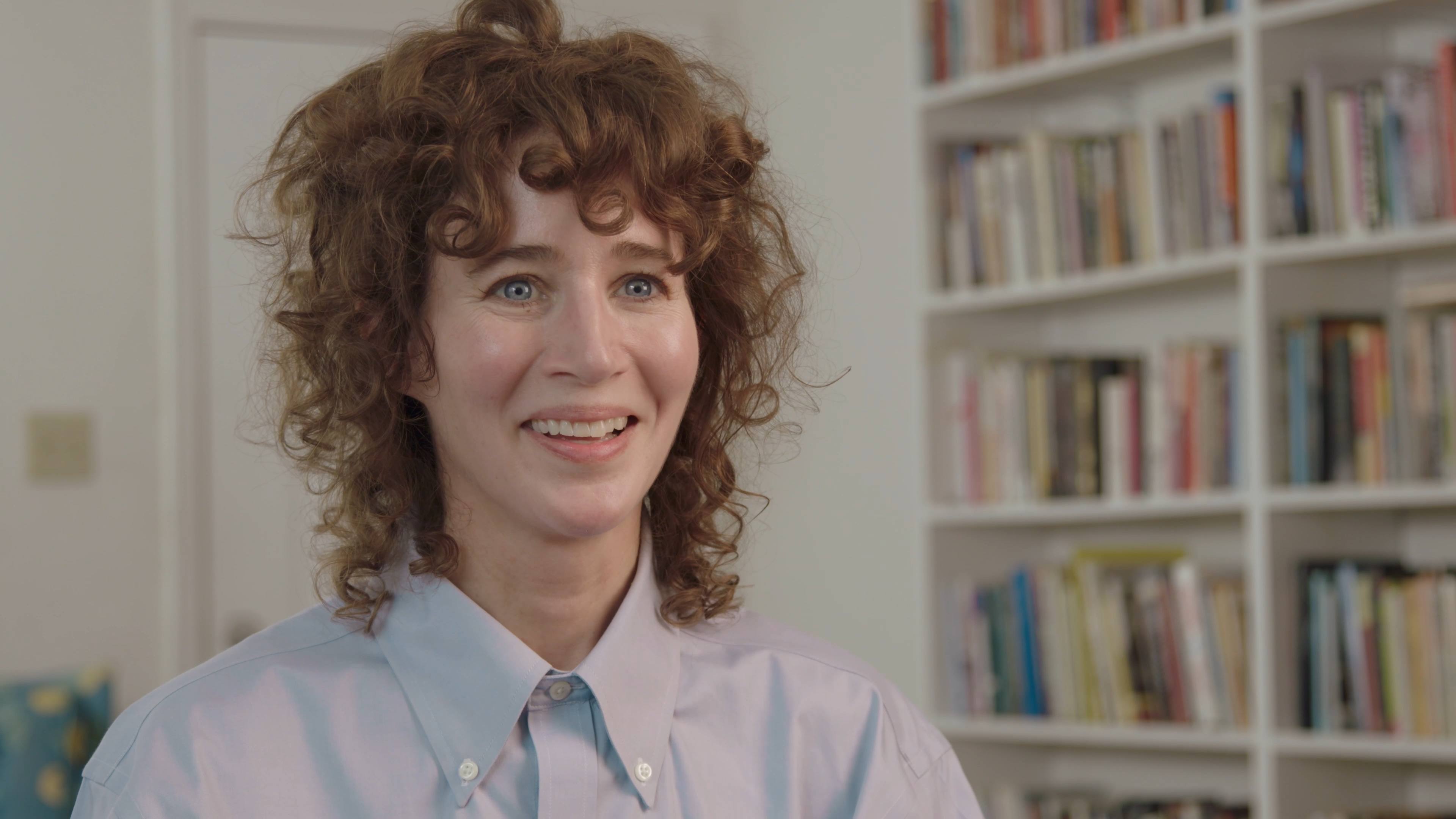 Miranda July with curly hair smiling, wearing a light blue button-up shirt. Bookshelves with various colored books are in the background.