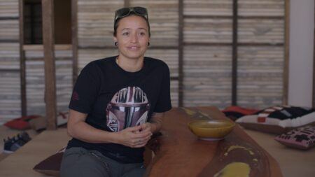 Rose B. Simpson hands clasped sitting in front of the wooden walls of her installation, "Dream House."