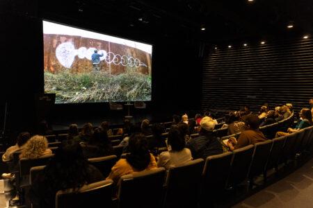Audience seated in a dark theater watches a large screen displaying street an artist spray painting on a rusted outdoor wall.