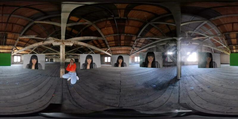 Three warped mirrors in an attic with five screens reflected showing the same woman with long black hair and blunt bangs. A person in an orange jacket and light wash jeans sits on the wooden floor to capture the scene.