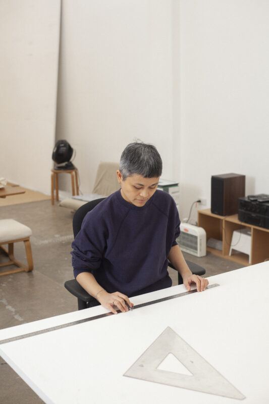 Maria Taniguchi sits at a desk, using a ruler and a triangular scale on a large white surface in a minimalist room.