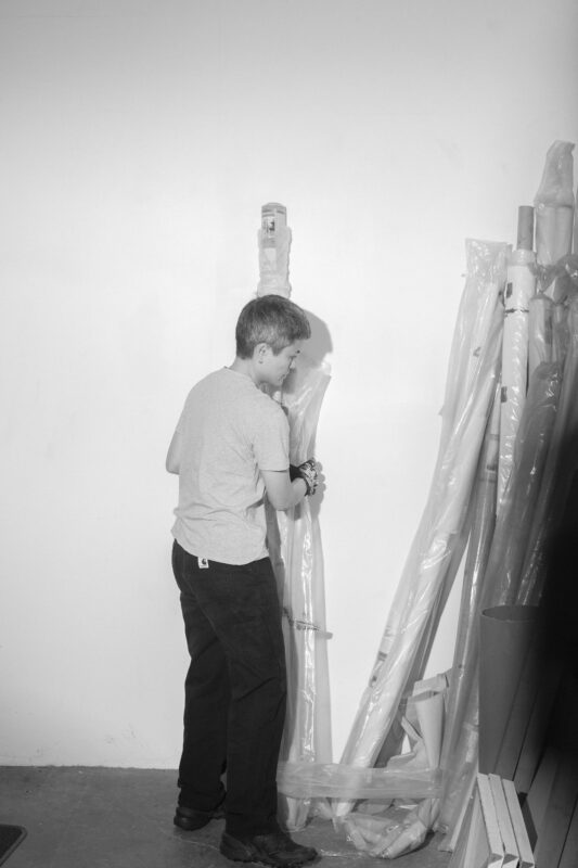 Black and White image of Maria Taniguchi holidng a long piece of material wrapped in plastic leaning against a wall.