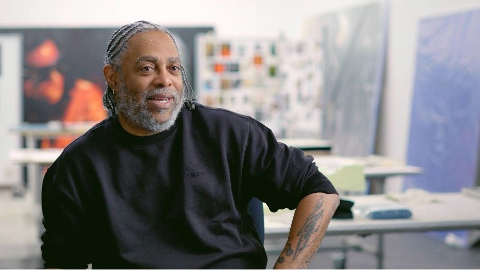 Arthur Jafa with gray braided hair and a beard sits in an art studio, wearing a black sweatshirt.