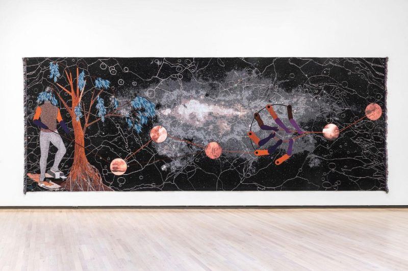 Otobong Nkanga's long black tapestry "Double Plot," a headless figure and tree stand to the left while orbs float in the middle. 
