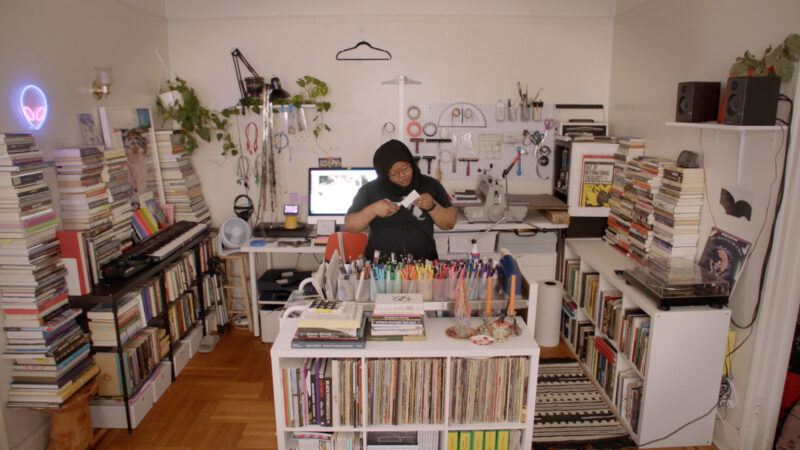 Kameelah Janan Rasheed in her Brooklyn studio filled with bookcases and stacks of books. She is cutting a piece of paper over a table filled with multicolored pens and markers. 