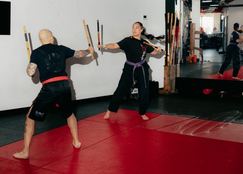 Two people practice stick fighting on red mats in a martial arts studio, facing each other with sticks raised. A mirrored wall reflects part of the scene.