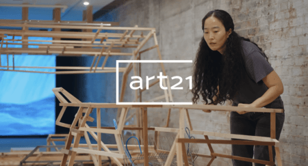 Hyeree Ro with long hair working on a wooden structure in cement room, with the Art21 logo overlaid on the image.