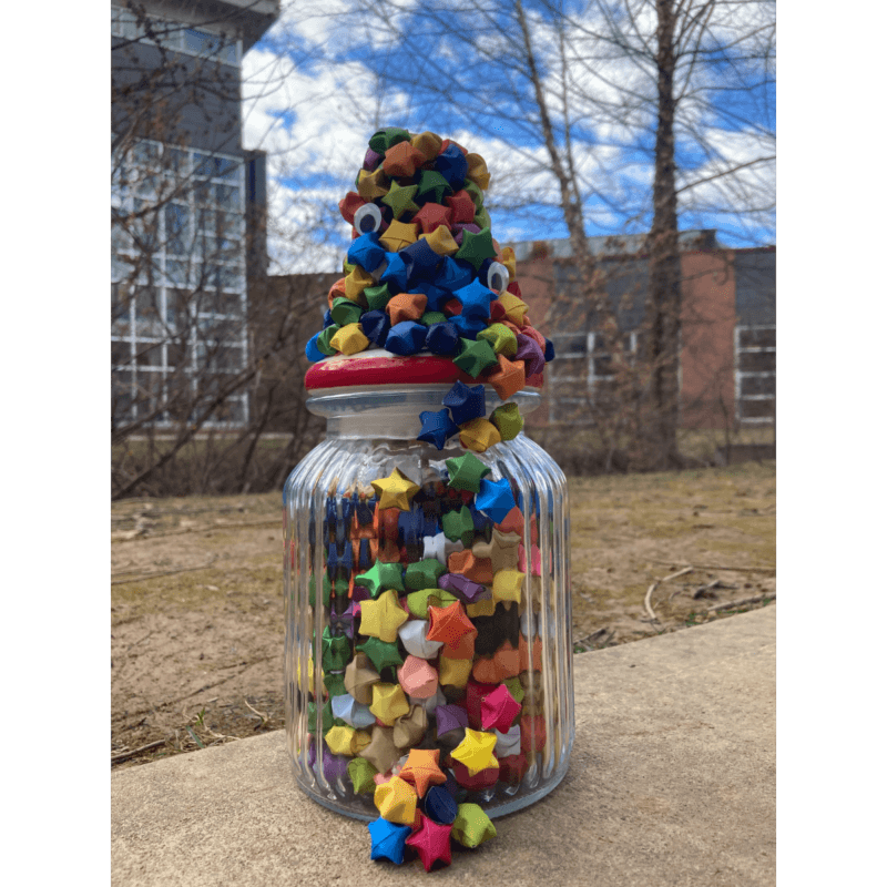 A glass jar filled with colorful paper stars sits outside on a concrete surface, stars with googly eyes are stacked and spilling out of the top.